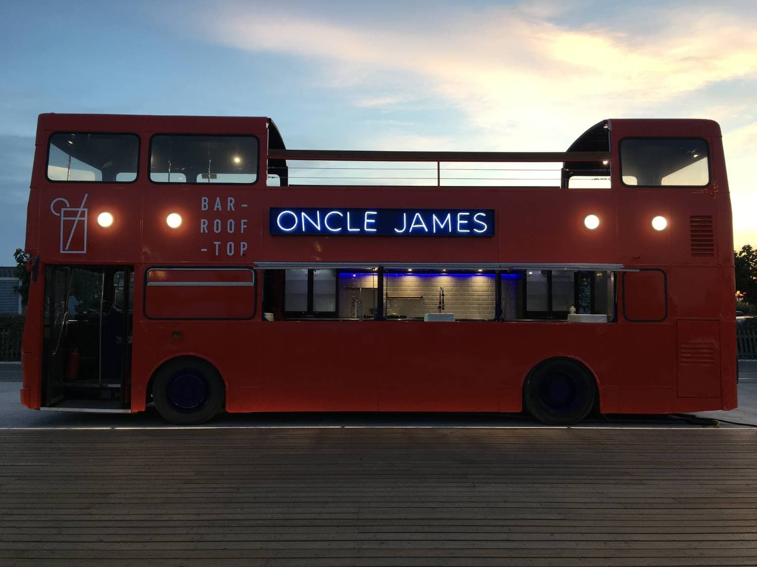 Double Decker rouge Oncle James avec enseigne néon bleue « Bar Rooftop » — bus bar événementiel au coucher du soleil